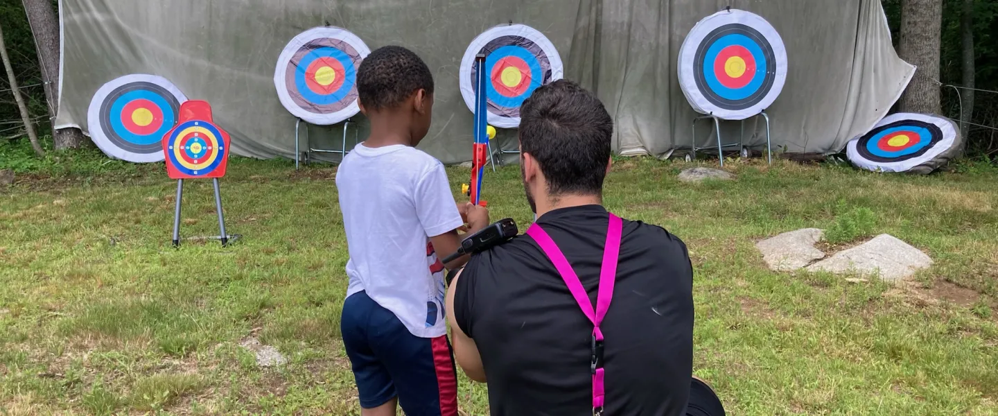 Male counselor assisting little boy with archery