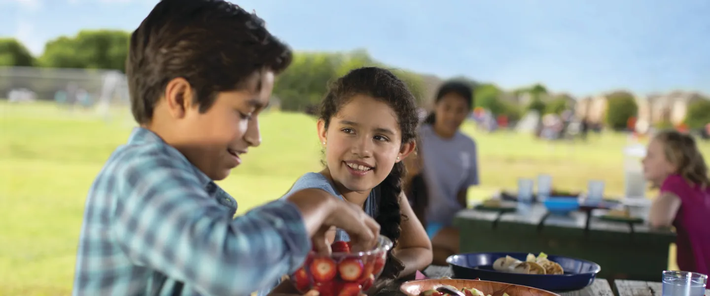 Boys and girls sitting at a picnic table outside eating food.