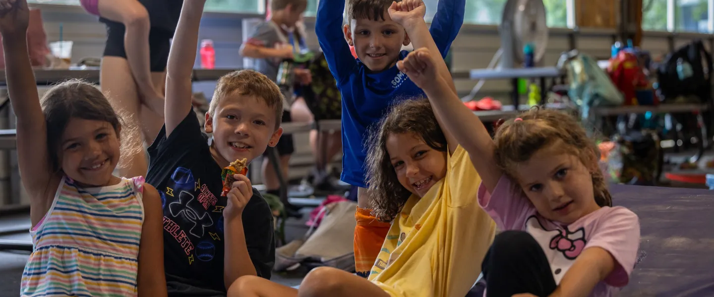 Four kids sitting on the ground raising their hands.