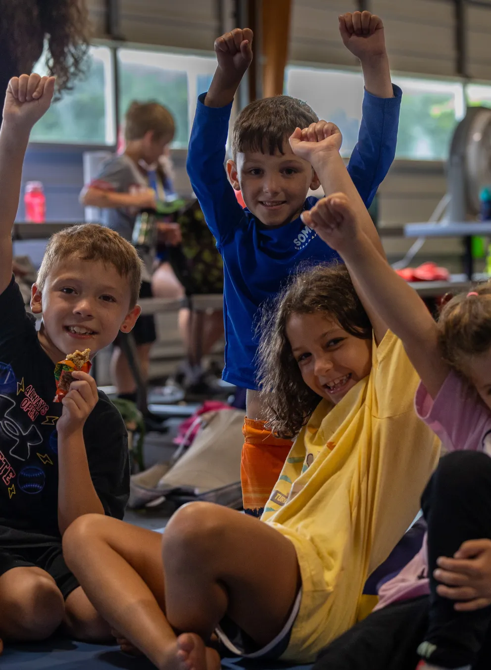 Four kids sitting on the ground raising their hands.