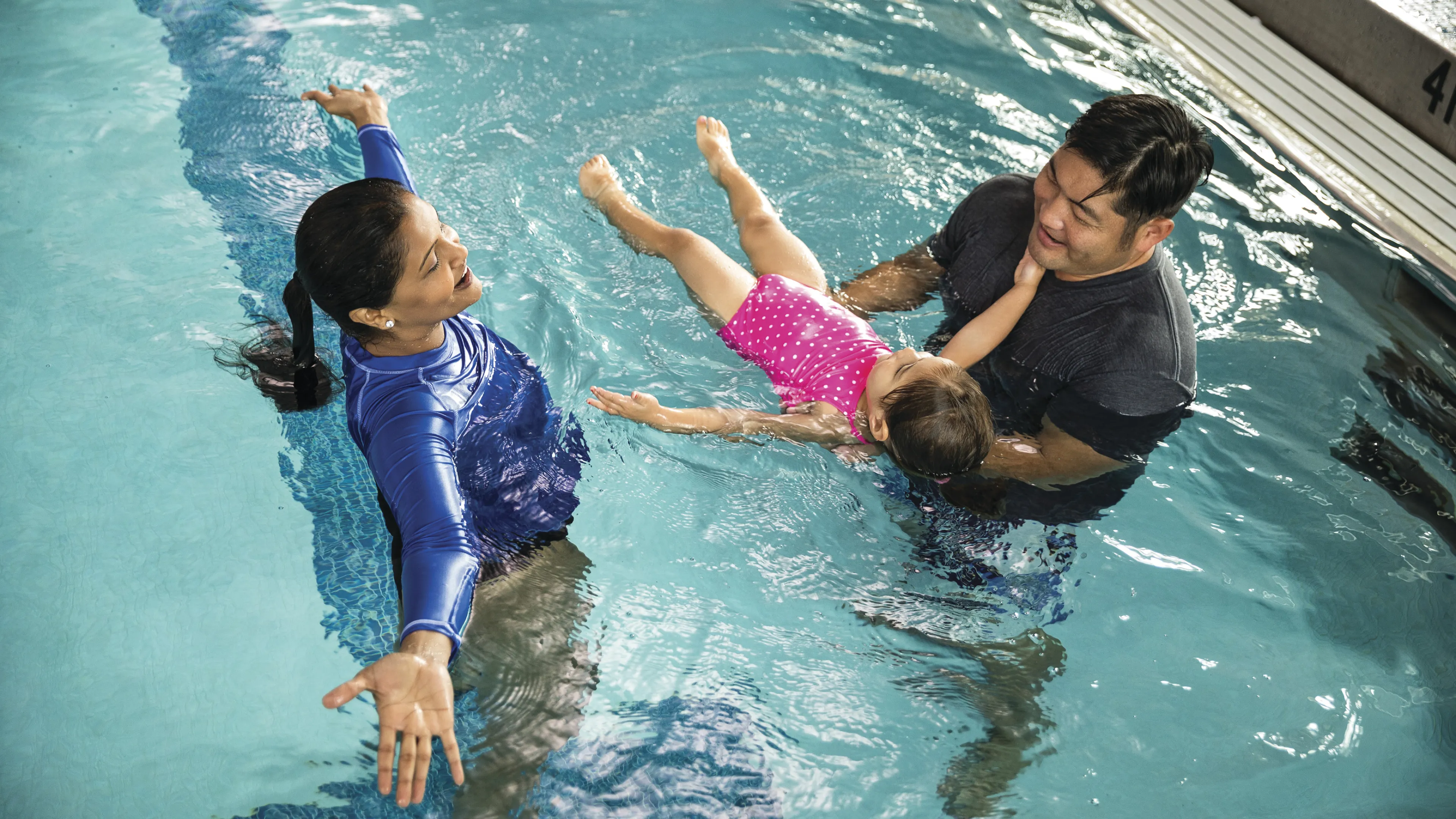 Man holding young girl on back in pool with female instructor