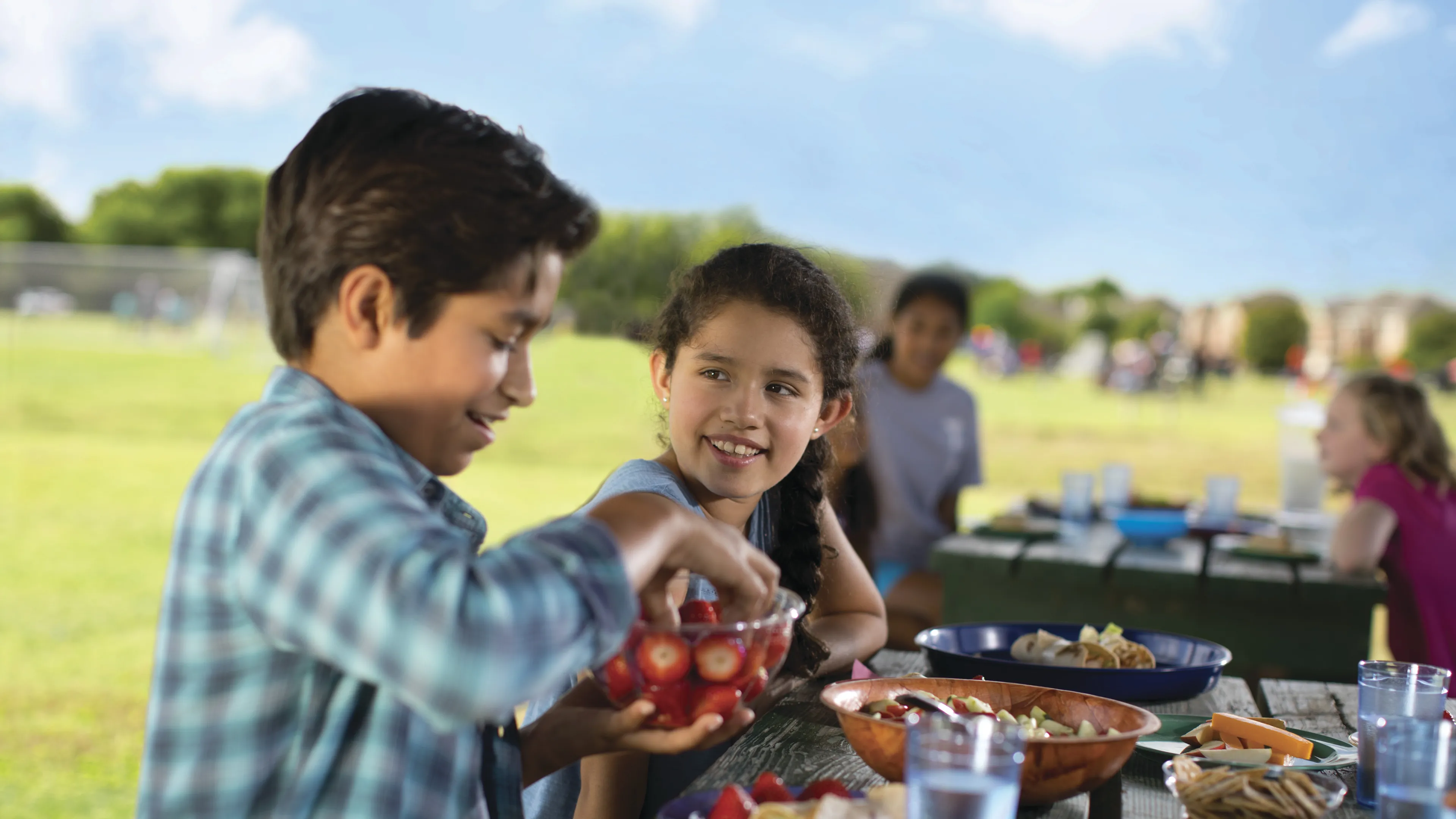 Boys and girls sitting at a picnic table outside eating food.