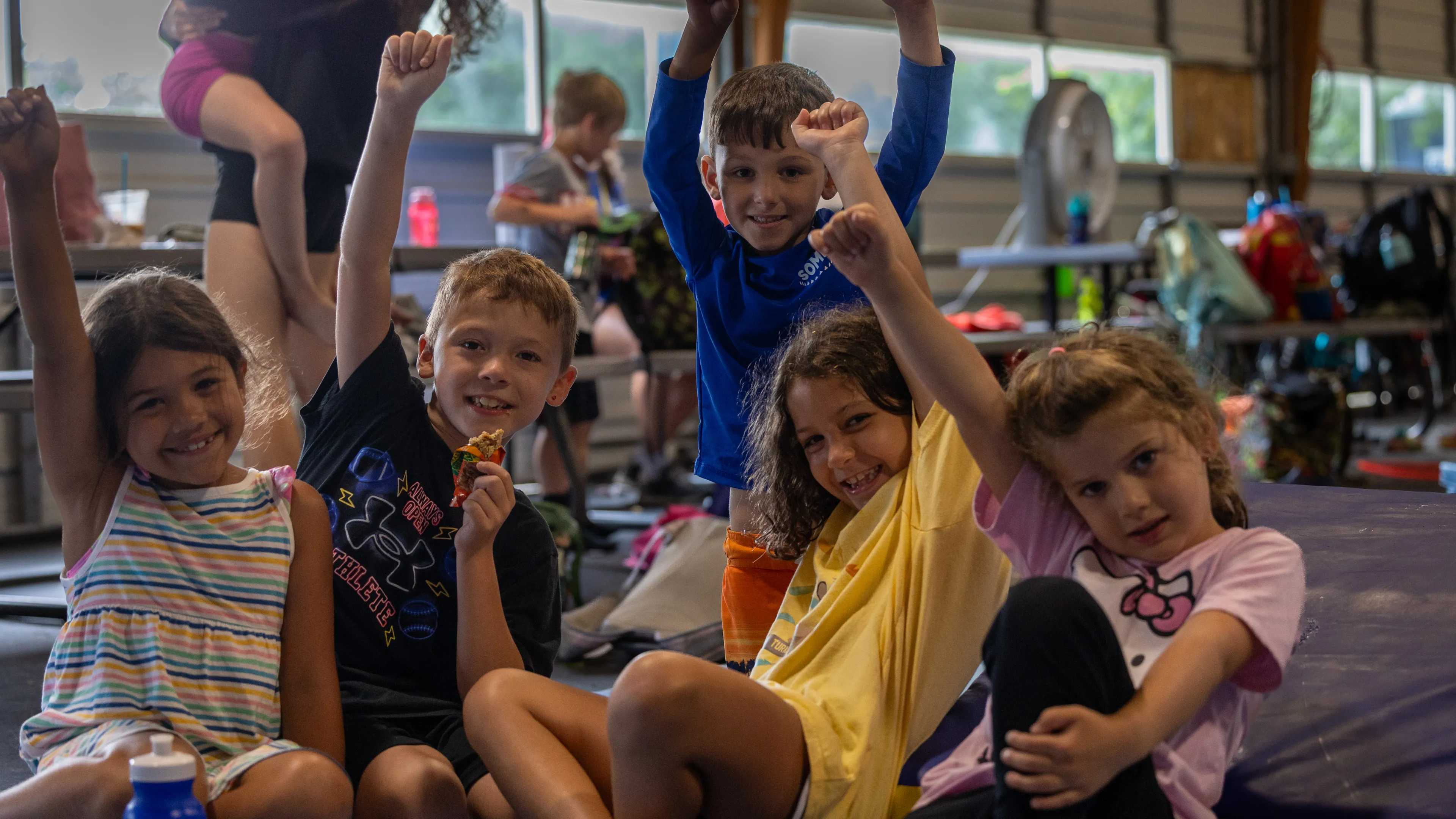 Four kids sitting on the ground raising their hands.