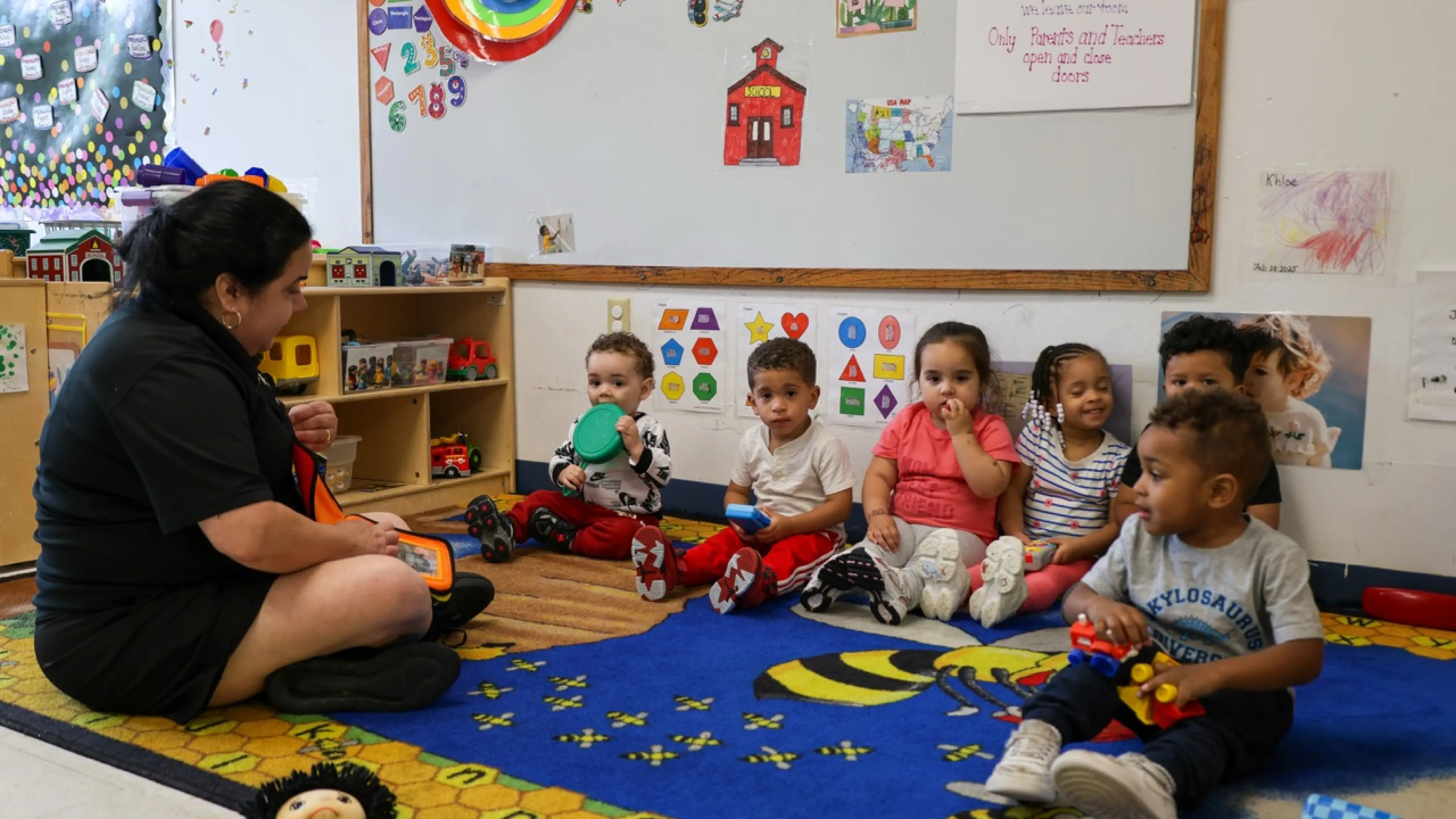 Group of six toddlers sitting on a rug infront of their teacher.
