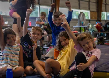 Four kids sitting on the ground raising their hands.
