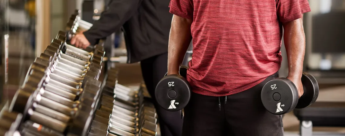Man holding dumbbells next to dumbbell rack with a female trainer behind him